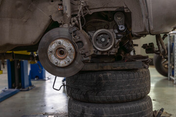 Close view of car brake disc and caliper reveals rusted hub, worn pads, dusty suspension hardware. Vehicle stands above stacked tires inside repair  © Egor Lint