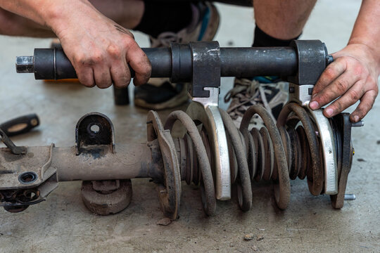 Hands work on car suspension repair, holding worn strut and coil spring parts on concrete floor. Dirty metal mounts, bolts, and rusted coils show