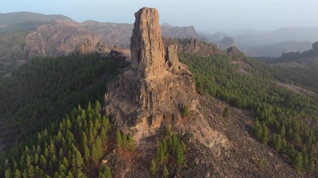 Majestic aerial view descending towards Roque Nublo, a volcanic rock formation in Gran Canaria, surrounded by a pine forest at sunset