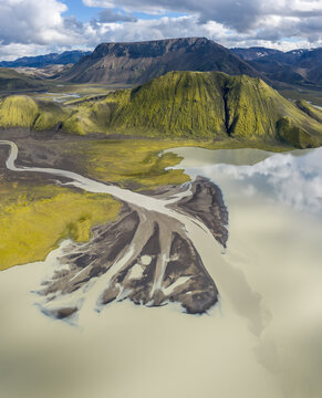 Aerial view of a glacial river delta flowing into a lake, surrounded by vibrant green moss-covered mountains and volcanic terrain under a cloudy sky in Iceland.