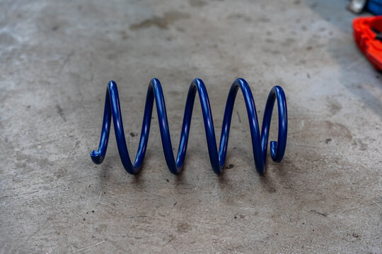 Single blue compression spring lies on rough concrete surface with scratched texture and stains. Industrial setting feels quiet, with soft background blur and red tool part at upper right.