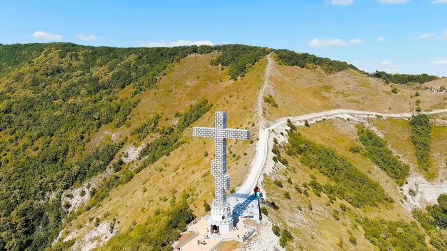 Gelendzhik, Russia. A towering white Orthodox worship cross with a lattice steel structure, aerial drone view against the forested slopes of the Caucasus Mountains under a clear summer sky. Drone foo