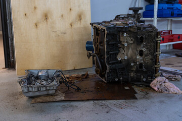 Broken vehicle engine block stands on stained floor beside open metal tray and loose parts. Grease marks, cloth rag, and workshop shelving create clear service bay setting for engine maintenance. © Egor Lint