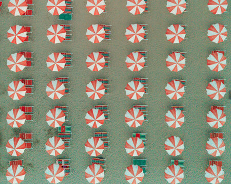 Aerial view of rows of red and white striped beach umbrellas and sun loungers arranged on the sandy beach in Cupra Marittima, Marche, Italy.