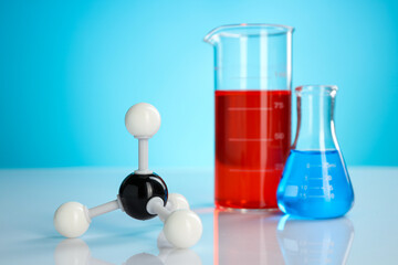 Chemical model of molecule and laboratory glassware on white table against light blue background,...