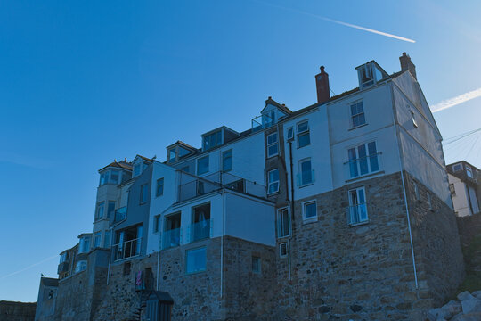 A large, multi-story building with a stone base and white upper floors, featuring balconies and dormer windows, stands against a bright blue sky - St Ives, Cornwall, UK
