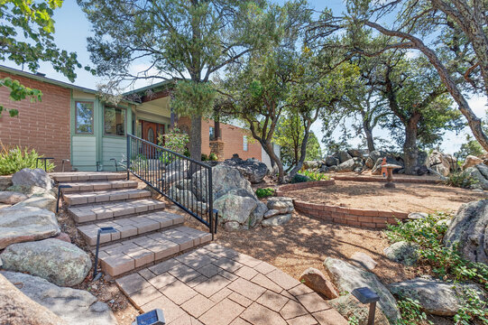 Brick house front yard with stone steps and iron railing, surrounded by trees and rocky garden entry