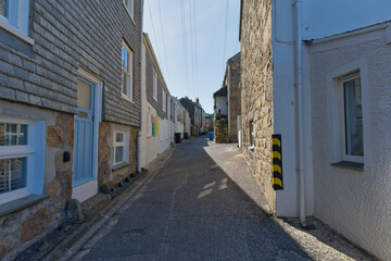 Fototapeta premium A narrow, sunlit street winds between stone and shingle buildings in a historic town. Overhead wires cross the bright sky - St Ives, Cornwall, UK