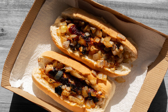 Hot dogs with fried onions in white finger bread rolls, made with pork sausages, in a cardboard takeaway tray background backdrop.