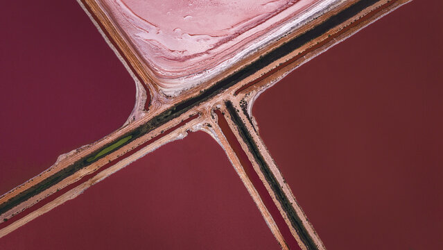 Aerial view of geometric salt evaporation ponds with vibrant pink and red water separated by dirt paths in Denham, Western Australia, Australia.