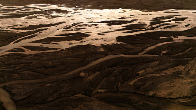 Aerial view of Kok Kiya Valley with its intricate braided river channels reflecting the golden light, surrounded by rugged mountain terrain in At-Bashi District, Naryn Region, Kyrgyzstan.