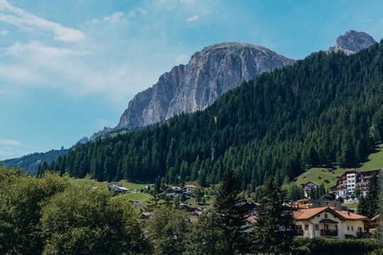 A Dolomite valley village nestles among pine forests below a rocky mountain ridge. Alpine resort town and summer mountain tourism for travel, holiday and destination marketing