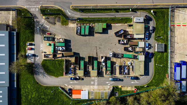 Aerial view of a waste recycling center with rows of green containers and bins, parked cars, and asphalt surfaces in Market Harborough, England, United Kingdom.