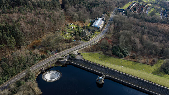 Aerial view of the Ladybower Reservoir featuring its iconic bellmouth spillway and dam, surrounded by dense woodland and rolling hills in the Peak District National Park, England, United Kingdom.