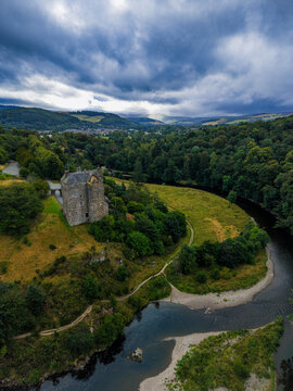 Aerial view of Neidpath Castle situated on the banks of the River Tweed surrounded by lush green hills and forests under a dramatic cloudy sky in Peebles, Scotland, United Kingdom.