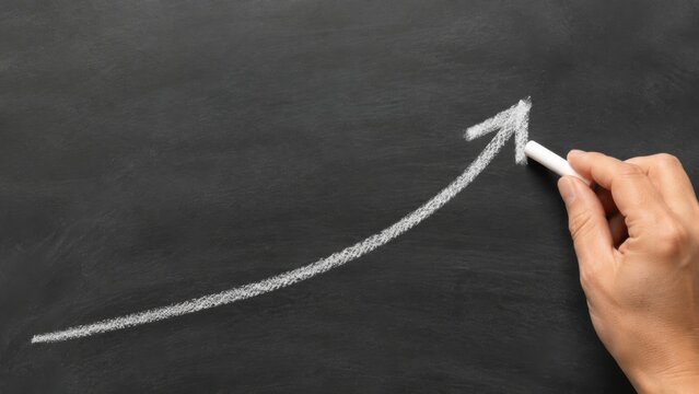 Hand drawing an upward arrow with white chalk on a black chalkboard surface, illustrating growth or progress in a clear and simple visual format