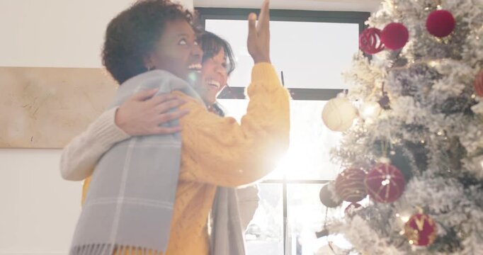 African American women in yellow, laughing by sunlit window hanging lit gold ornament on tree