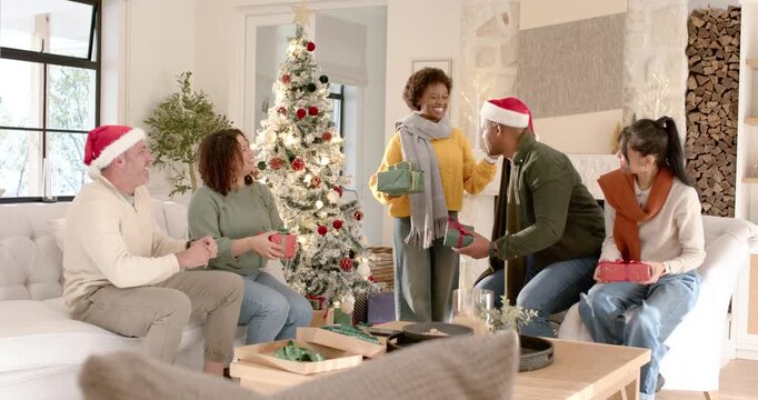 Diverse friends exchanging gifts by Christmas tree watching man in Santa hat offering present