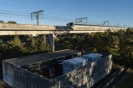 Aerial view of a metro traction substation with a train on an elevated track, surrounded by trees under a clear sky in Rouse Hill, Sydney, New South Wales, Australia.