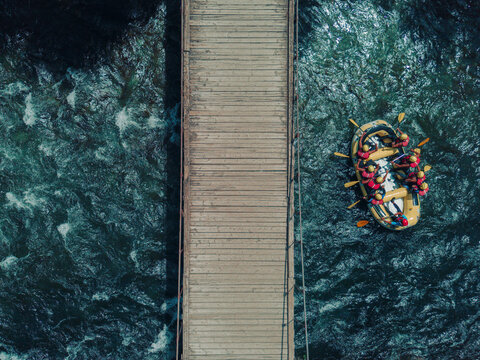 Aerial view of a wooden footbridge spanning the Noguera Pallaresa river with a yellow raft carrying people in life jackets and helmets navigating the rapids in Lleida, Catalonia, Spain.