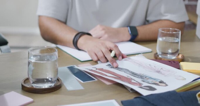 Designer is pointing sketches at wooden table as peer is placing new sheets while selecting designs