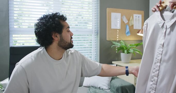 Man sitting on bed turning to off-frame hand holding pink shirt, inspecting drape for fit
