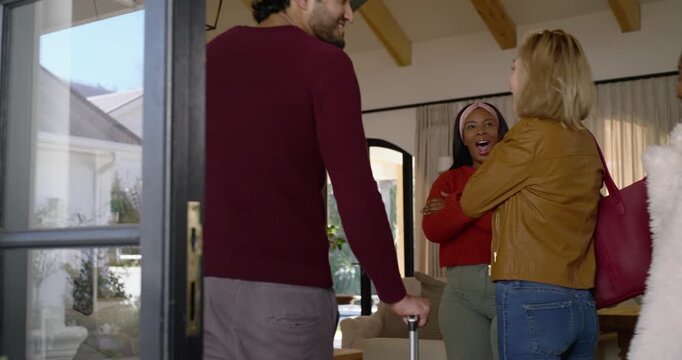 Diverse friends entering living room via glass door as host greeting, man with suitcase for holiday
