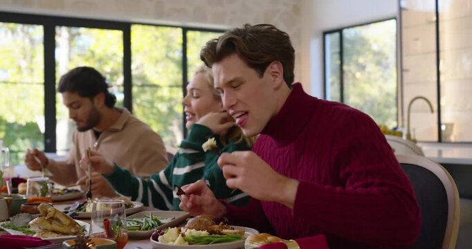 Diverse friends in sweaters at table, plates being set out, eating, toasting glasses to celebrate