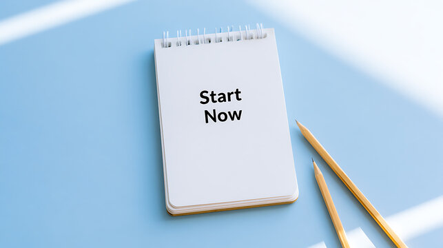 A blank notebook with the message start now written on the white paper next to two yellow pencils on a clean blue office table surface