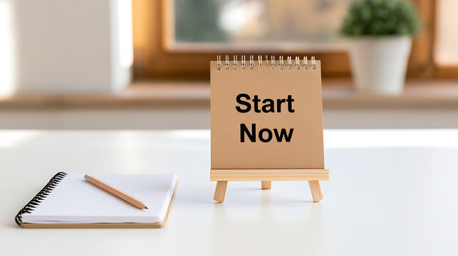 Inspirational start now message written on a paper stand on an office desk with a notebook and pencil for business motivation goals and success