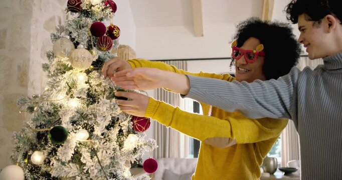 African American male friends placing ornament on flocked Christmas tree at home after bauble pass