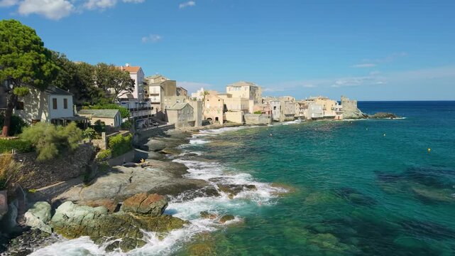 Erbalunga Corsica drone flyover along coastal rocks Genoese tower emerald sea waves crashing traditional seaside stone houses solitary people on boulders Cap Corse wild landscape