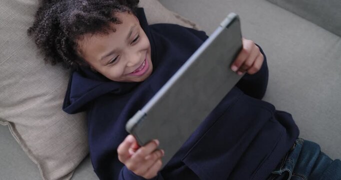 African American child on couch with pillow holding tablet reacting to screen and adjusting grip