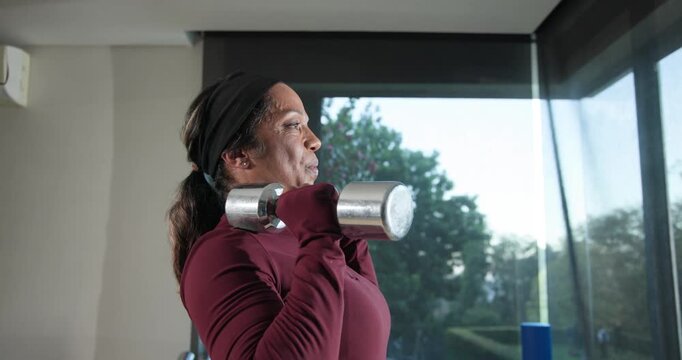 African woman in maroon starting front rack pressing chrome dumbbells alternately by tall window