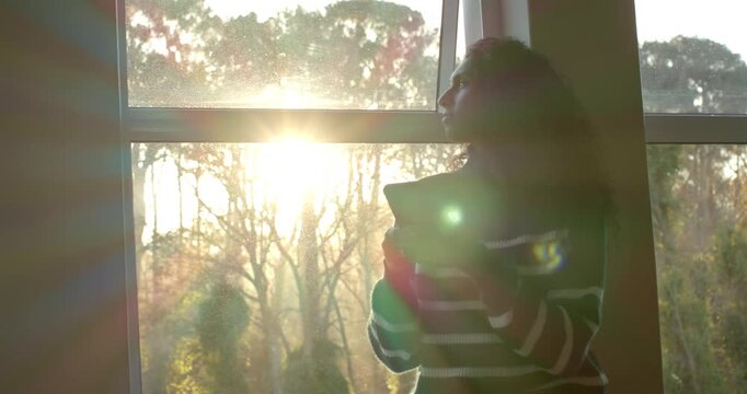African woman in striped sweater holding device by window panes, sun rising causing gaze
