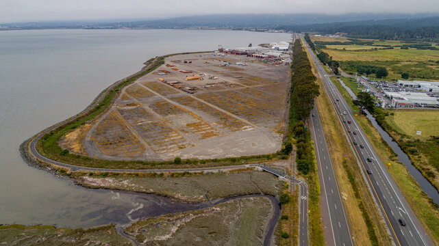 Aerial view of a wood mill industrial site located along the waterfront, featuring large open gravel areas, nearby highway traffic, and coastal marshland in Eureka, California, United States.