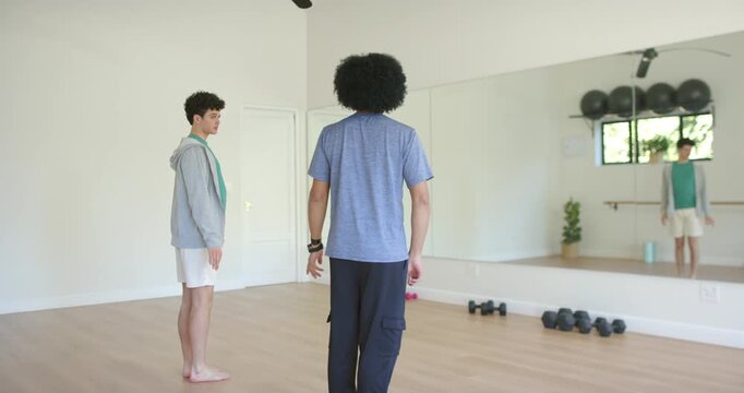 Diverse male coach guiding student by raising hands, both holding tree pose by mirror, copy space