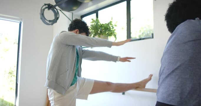 Diverse male duo stretching at bright studio, starting with left leg on barre, practicing balance