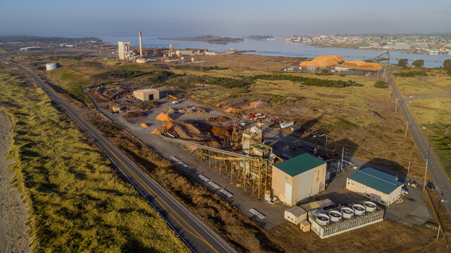 Aerial view of the industrial pulp mill facility featuring large piles of wood chips, processing machinery, and storage buildings near the coastline in Samoa, California, United States.