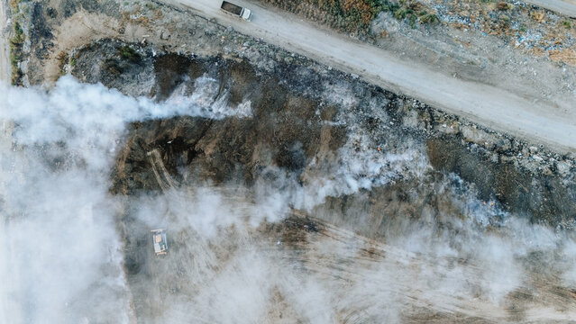 Aerial view of a landfill site with smoke rising from burning waste, heavy machinery operating on the ground, and dirt roads in Sremska Mitrovica, Vojvodina, Serbia.