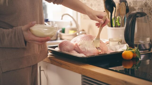 Festive holiday meal preparation with woman carefully basting a large raw turkey on a baking sheet, using a brush to spread a savory sauce before roasting it in the oven for dinner