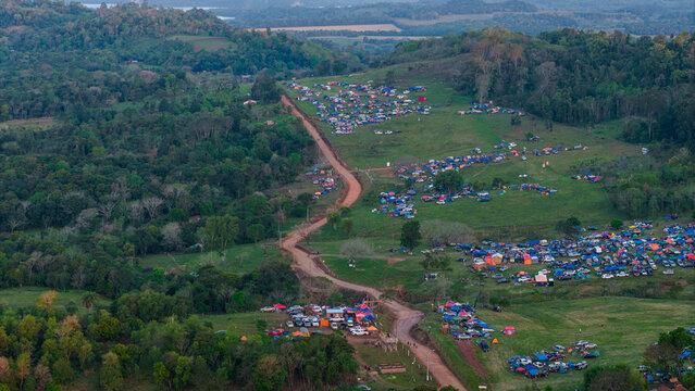 Aerial view of Rally Paraguay 2026 event with colorful tents and vehicles along a winding dirt road through the green hills and forests of Nueva Alborada, Itapua Department, Paraguay.
