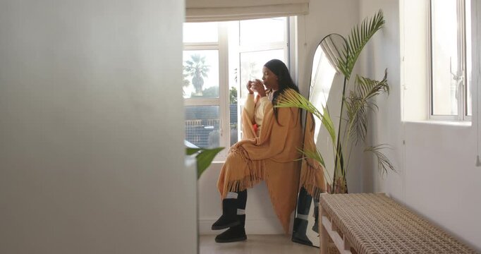 Adult African American woman in shawl holding mug at window, sipping while gazing at palm outside