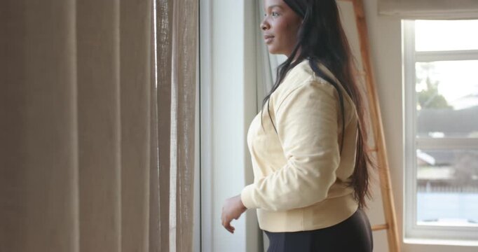 African American adult woman pulling curtain aside at sliding door, scanning view, checking phone