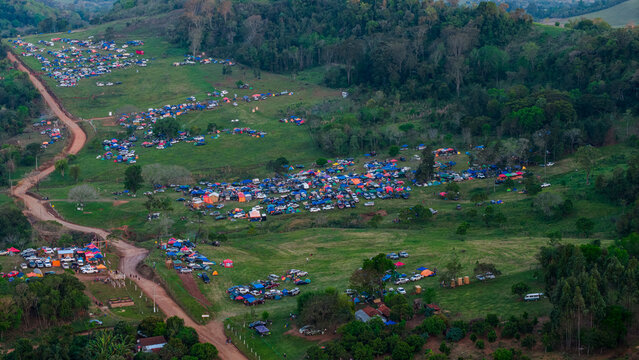 Aerial view of Rally Paraguay 2026 campsite with numerous tents and vehicles on a green hillside, featuring dirt roads and forest, Nueva Alborada, Itapua Department, Paraguay.