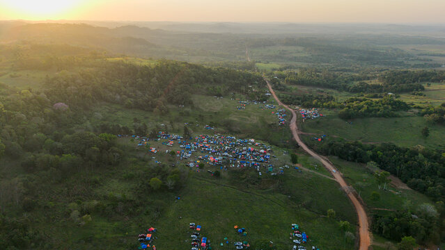 Aerial view of Rally Paraguay 2026 campsite with numerous tents and vehicles scattered across a rural hilly landscape during golden hour in Nueva Alborada, Itapua Department, Paraguay.