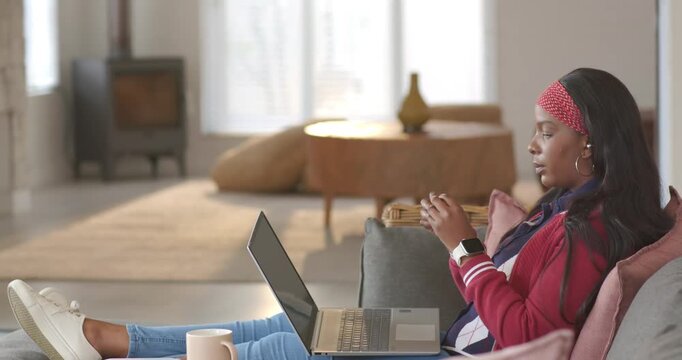 African American woman checking laptop, inserting earbud for audio and adjusting on sofa