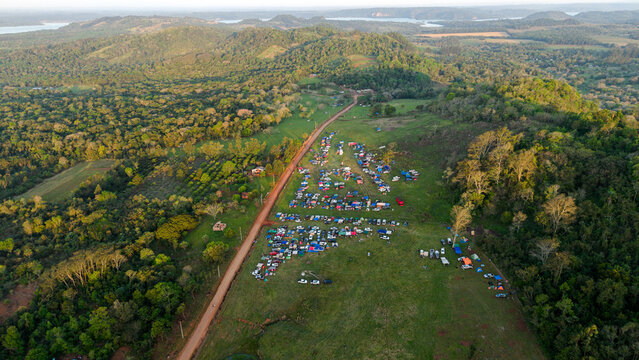 Aerial view of Rally Paraguay 2026 campsite with vehicles and tents on a grassy field beside a dirt road in a hilly, forested landscape in Nueva Alborada, Itapua Department, Paraguay.
