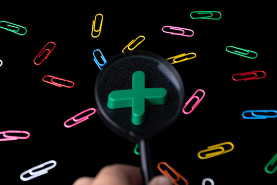 Hand holding magnifying glass over green plus symbol and colorful paper clips on black background