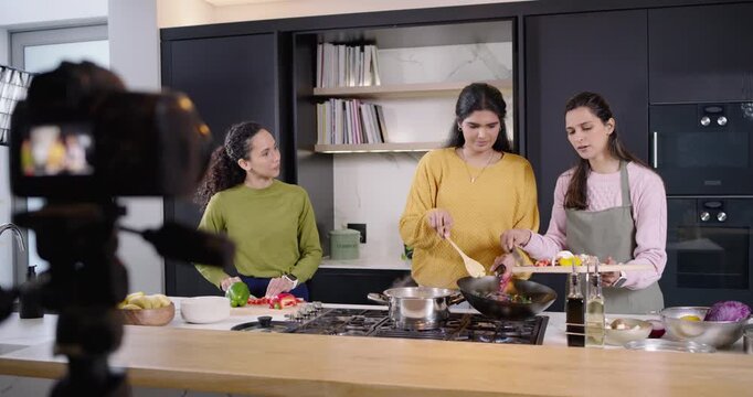 3 women, one wearing apron, chopping, stirring wok at kitchen island with camera light filming demo